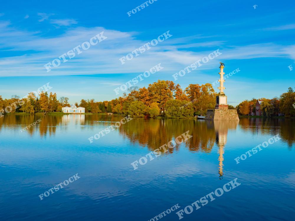На фотографии изображена Чесменская колонна, расположенная на Большом пруду в Екатерининском парке Царского Села. Этот монумент был воздвигнут в честь победы России в Чесменском сражении. Вокруг колонны раскинулись осенние деревья, а спокойная вода пруда отражает её и окружающую природу.